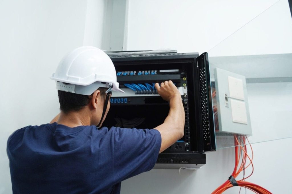Technician in a hard hat wiring an open wall-mounted patch panel with blue cables